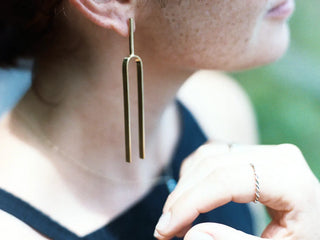 A close-up lifestyle shot of a person wearing a solid brass tuning fork earring. The earring hangs from a simple post and features two parallel vertical bars.